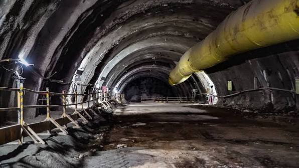 Inside an unfinished excavated tunnel with a yellow ventilation duct and safety railings along the wall