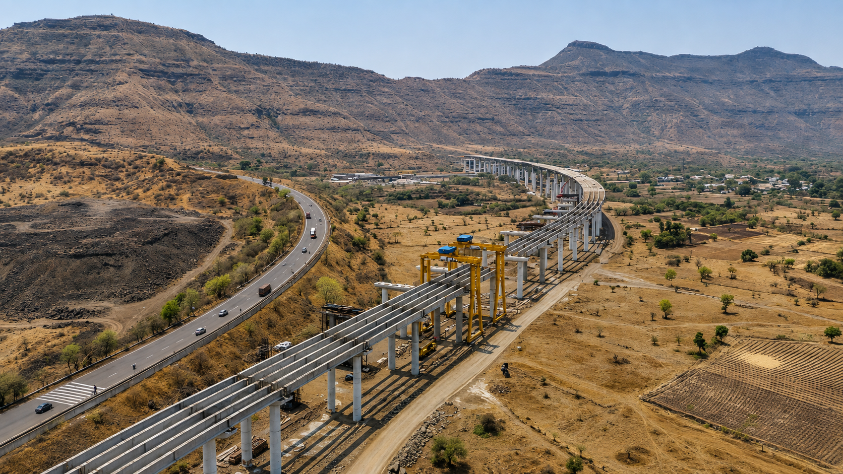 Aerial view of an elevated viaduct under construction curving beside a highway through arid hills